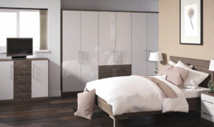 Bedroom with a modern design. Features a large bed with white and beige bedding, built-in wardrobe, and a dresser with a TV. Theres a lamp on a nightstand and a framed floral picture on a textured wall. Dark wood flooring completes the look.