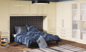 A modern bedroom features a bed with a blue-striped duvet and multiple pillows. Its surrounded by cream-colored built-in wardrobes and cabinets. A stack of books sits on the wooden floor, and a circular black ceiling light hangs above.