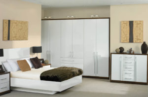 A modern bedroom featuring a white bed with brown and tan pillows, a dark fur throw, and matching side tables with black lamps. White wardrobes and a dresser complement the decor, with abstract wooden art pieces on the walls.