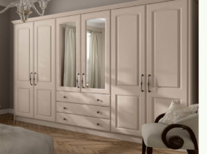 A beige wardrobe with multiple doors and drawers, featuring mirrored panels, is situated against a wall in a bedroom. The wardrobe has black handles, and a window with light curtains is partially visible. A chair with a cushion is placed nearby.