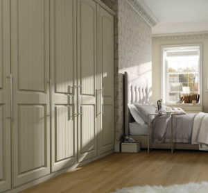 A cozy bedroom with a large bed featuring a tufted headboard and gray bedding. Sunlight filters through a window, illuminating the room. There are built-in wardrobes on one side and a fluffy rug on the wooden floor.
