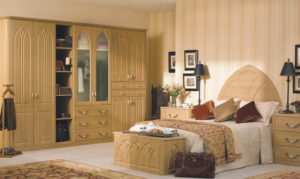 A cozy bedroom with a wooden wardrobe featuring arched details, a bed with a decorative headboard, and a matching chest at the foot of the bed. The room has warm lighting, framed art on the walls, and a mix of pillows and blankets.