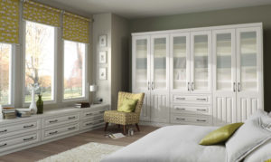 A bright bedroom with large windows, white cabinets, and a bed with green and white bedding. A chair with a yellow patterned fabric sits near the window. The walls are light green, and the room has a soft, neutral-toned color palette.