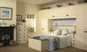 A cozy bedroom with cream-colored furniture, featuring a bed with blue and white bedding. Shelves and cabinets surround the bed. A window with striped curtains, a chest at the foot of the bed, and a fireplace add warmth to the room. A picture hangs on the wall.
