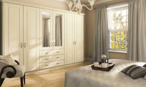A cozy bedroom featuring a bed with a grey patterned bedspread and a tray with a beverage set. Cream-colored wardrobes with mirror panels line the wall, and a window with grey curtains lets in natural light. A chandelier hangs from the ceiling.