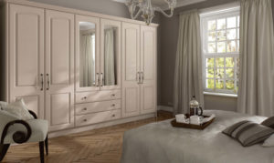 A cozy bedroom with beige wardrobes and mirrored cabinet doors. Theres a bed with a patterned duvet and decorative pillows. A tray with a teapot and cups is on the bed. Large window with curtains allows natural light in, and a chandelier hangs from the ceiling.
