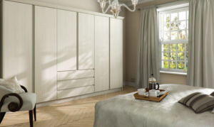 A cozy bedroom with a light wood wardrobe, a large window with light curtains, and a bed covered in a gray patterned duvet. A tray with a French press and cups is on the bed. The room features a herringbone wood floor and a cushioned chair.