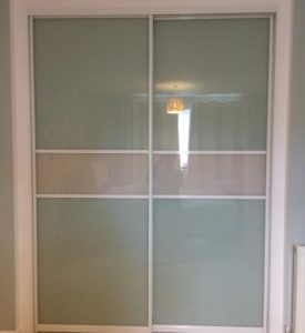 Sliding closet doors with a frosted glass finish in a light, neutral room. The doors have two vertical panels with a horizontal opaque stripe across the middle. A small chandelier is reflected in the glass.