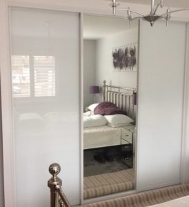 A bedroom with a metal bed frame, white bedding, and purple pillows. A mirrored closet reflects the bed. A chandelier hangs from the ceiling, and there is a window with blinds on the left side.