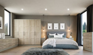 A modern bedroom with gray walls, featuring a large bed with pillows and a throw blanket. Two pendant lights hang above nightstands on either side. A textured rug lies on the floor, and three framed artworks are displayed above the headboard.