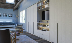 A modern bedroom features a large wardrobe with built-in shelves and a TV. Two sleek chairs and a small table sit on a cozy rug. The room has wooden flooring, light blue walls, and a skylight. Black and white photos decorate the wall near the bed.