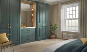 A bedroom with teal built-in wardrobes and drawers, featuring a large mirror and wooden accents. A window with white curtains lets in natural light. Theres a bed with teal and mustard bedding and a potted plant nearby.