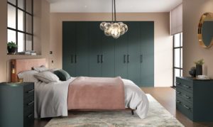 Modern bedroom with a green color scheme featuring a large bed with pink bedding, flanked by green nightstands. A cluster of pendant lights hangs above. There are green wardrobes and a large window with soft natural light filling the room.