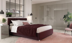 A modern bedroom with a burgundy bed, white and pink bedding, and decorative pillows. Two pendant lights hang over bedside tables with books and lamps. A large mirrored wardrobe and indoor plant complete the rooms decor.