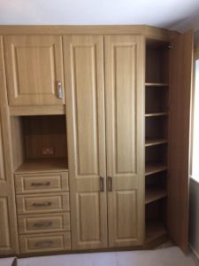 Wooden corner wardrobe with multiple compartments. It features a section with drawers, a tall unit with a closed cabinet, and an open shelving unit on the right. Natural light from a nearby window partially illuminates the furniture.