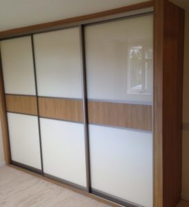 A large wardrobe with sliding doors featuring a modern design. The doors have a combination of white and light wood panels. The wardrobe is placed against a beige wall and theres a window in the background reflecting light.