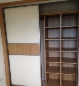 A modern closet with sliding doors, one made of white panel with a wooden accent and the other open, revealing a series of wooden shelves. The closet has a simple and clean design.