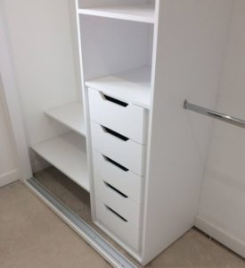 A walk-in closet with white shelves and five drawers on the right side, an empty rail for hanging clothes, and beige flooring.