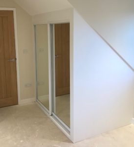A small interior space under stairs with beige walls and light brown wooden doors. The area features mirrored sliding closet doors, reflecting parts of the room. The floor is light-colored, either carpeted or unfinished.