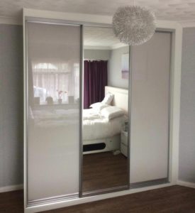 A bedroom reflected in a large sliding mirror wardrobe. The reflection shows a neatly made bed with white bedding, a bedside table, and a window with sheer and dark curtains. A modern, textured light fixture hangs above.