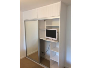 A white wardrobe with sliding mirrored doors and open shelves is shown. One shelf holds a small flat-screen TV, with a power cord leading to a socket on the beige carpeted floor. The wardrobe is set against a plain wall.