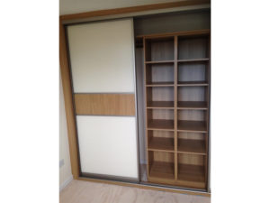 A wooden closet with sliding doors. One door is open, revealing shelves on the right side with multiple compartments. The left door is closed, featuring a white panel with a wooden strip across the middle.