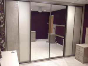 A spacious bedroom with dark purple walls and carpeted floor. The room features large mirrored sliding closet doors, a ceiling fan, and matching beige dressers on either side. A small remote is on the dresser.