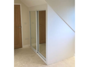 The image shows an interior corner of a room with a sloped ceiling. A wooden door is on the left side, and opposite the door are built-in sliding mirrored closet doors. The floor appears to be light-colored carpet.