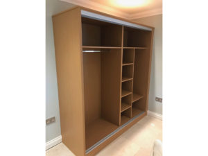 A large wooden wardrobe with multiple open compartments, including a section with a clothing rail and several shelves for storage, is set against a light-colored wall in a room with a tiled floor.