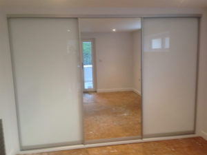 A room with sliding glass doors featuring frosted glass panels. The doors are partially open, leading to another space with a window and a small door. The floor is bare with a plywood surface. Walls and ceiling are painted white.