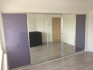A room with light gray wood flooring and a three-panel sliding wardrobe. Two panels are mirrored, and one is opaque with a purple finish. A closed wooden door is visible in the background.