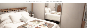 A cozy bedroom with a wooden bed, white bedding, and a decorative quilt. There are mirrored wardrobe doors reflecting the room. The wall displays the text Chichester Bedrooms at the top.
