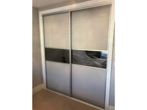 A sliding closet door with a modern design featuring a frosted white upper and lower panel, divided by a horizontal section of reflective glass, set within a silver metal frame. The closet is in a carpeted room with light-colored walls.