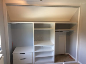 Custom-built white closet with open shelving and drawers under a sloped ceiling. It features a small door on the right side and an unfinished section, suggesting ongoing work. The interior is bright with ample storage space.