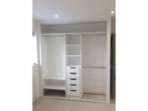 A built-in white closet with open sliding doors. It features shelves, a set of drawers, and multiple hanging rods, providing a spacious storage solution in a minimalistic room with white walls and a light floor.