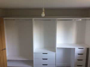 A white, built-in wardrobe with several open compartments and drawers. An empty hanging rod is visible under a bare light bulb on the ceiling. The wardrobe is positioned against a blue-gray wall and a wooden door is partially visible on the left.