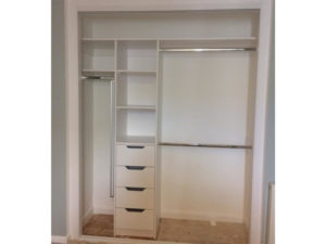 An empty white open closet with multiple sections, including shelves, four drawers, and two metal hanging rods. The closet interior is surrounded by a light-colored wall, with wooden flooring visible at the bottom.