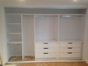 A built-in white wardrobe with open sections. It features shelves on the left, a center space for hanging clothes, and two sections with drawers on the right. The floor is bare, suggesting its in a newly constructed or renovated room.