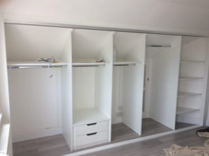 A large empty white wardrobe with open sliding doors, featuring hanging rods, shelves, and drawers. The room has laminate flooring, and a pile of beige laundry is on the floor to the right.