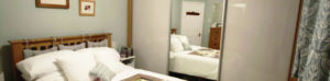 A cozy bedroom with a wooden bed, white bedding, and decorative pillows. A large mirrored wardrobe reflects the room. Framed pictures hang on the walls, and a partially visible door leads to another room. Patterned curtains adorn the window.