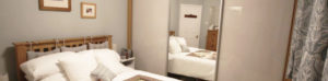 A cozy bedroom with a wooden bed featuring white bedding and pillows. A large mirror reflects the room, including framed pictures on the wall, a dresser, and patterned curtains. The walls are painted a soft gray.