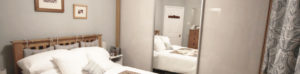 A cozy bedroom with a wooden bed, white bedding, and decorative pillows. Large mirrored wardrobe doors reflect the bed and a dresser with a closed door in the background. The room features framed pictures on a light gray wall and patterned curtains.