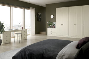 A spacious, modern bedroom with light wood flooring, white wardrobes, and a large window with sheer curtains. Theres a bed with dark bedding, a desk with a chair, potted plants, and a wall clock. The walls are painted dark green.