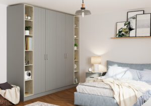 A cozy bedroom features a large wardrobe with vertical panel doors and open shelves displaying books and decor. A modern pendant light hangs above a bed with striped bedding, and framed art rests on a shelf above a lit bedside table.