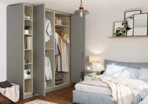 A cozy bedroom with an open corner wardrobe displaying clothes and books. A plush bed with striped linens is beside a nightstand holding a lamp. Art frames and a plant adorn a small shelf above the bed. A single pendant light hangs from the ceiling.