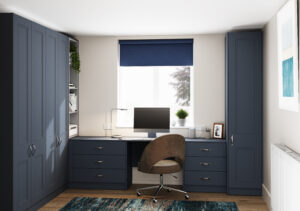 A modern home office setup featuring a dark blue L-shaped desk with cabinets and drawers. A computer monitor sits on the desk, with a brown swivel chair in front. A window with a blue roller blind lets in light, and the walls are adorned with a framed print.