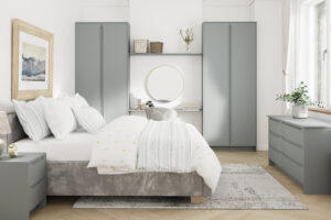A modern bedroom with a gray and white color scheme. Features include a neatly made bed, gray cabinets, a round mirror, and a dresser. A framed artwork hangs on the wall, and a large window on the right lets in natural light.
