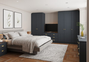 A modern bedroom features a plush bed with white linens and a gray headboard. Dark blue wardrobes and a dresser provide storage. A TV sits on the dresser. Two framed botanical prints hang above the bed. A patterned rug covers the wooden floor.