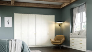 A cozy bedroom features a built-in wardrobe with cream-colored doors, an armchair, and a window seat beneath a bay window with curtains. The room has a dark hardwood floor and exposed wooden beams on the ceiling.