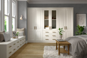 A cozy bedroom with a large white wardrobe featuring glass panels and drawers. Theres a cushioned window seat with pillows on the left, a soft rug on the wooden floor, a small wooden table, and a bed. Plants and minimal decor complete the space.